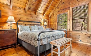 Ein Schlafzimmer einer Holzhütte mit einem Bett, Nachttischlampen, einer Bank und einem Fenster mit Jalousien.