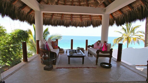 A covered outdoor seating area with a view of the ocean, furnished with chairs and a table, surrounded by palm trees.