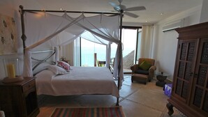 A bedroom with a four-poster bed, a ceiling fan, a view of the sea, a wooden cabinet, and a striped rug.