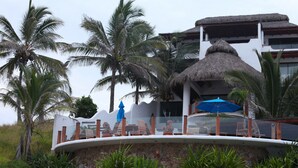 A resort with a thatched-roof structure, palm trees, and a pool area with lounge chairs and umbrellas.