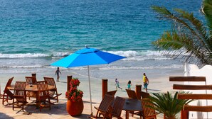 A beachside patio with wooden chairs and tables, a blue umbrella, and a view of the ocean.