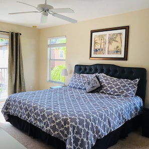 Quarto com cama, ventilador de teto, janela com cortinas e um quadro na parede.