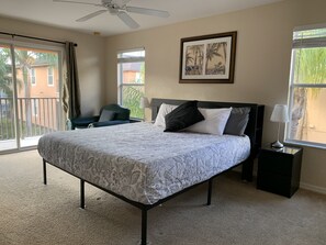 A bedroom with a bed, a chair, a nightstand, a ceiling fan, and a window with a view of palm trees and buildings. - Lovely 2 King Size Ensuite BR , 3 Mi to Disney, 1min to Pool, Free WiFi &Parking (Kissimmee)