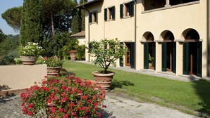 A building with a beige facade, green shutters, and a garden with potted plants and flowers.