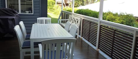 Eine überdachte Terrasse im Freien mit weißem Tisch und Stühlen, einem Grill und Blick auf die umliegende Grünfläche.