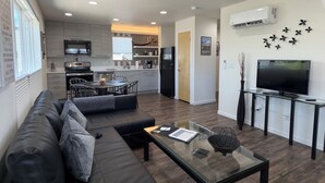 Ein modernes Wohnzimmer mit Sofa, Couchtisch und Fernseher. Im Hintergrund ist eine Küche mit Essbereich zu sehen.