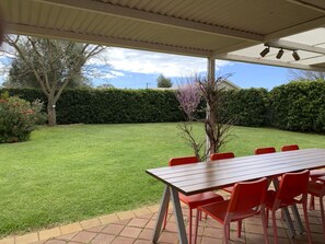 A covered patio with a wooden table and red chairs, surrounded by a well-maintained lawn and a hedge.