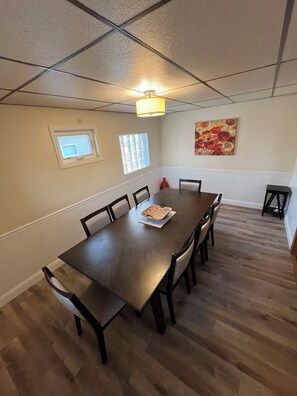 Une salle à manger avec une table rectangulaire, six chaises, un luminaire au plafond, une fenêtre et un tableau accroché au mur.