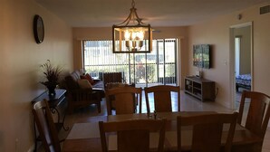 A spacious living room with a dining area, large windows, a chandelier, and a television on the wall. - Cozy and Charming Condo in Naples (Naples)