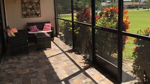 A sunlit balcony with wicker furniture, a ceiling fan, and a view of a well-maintained lawn and palm trees. - Cozy and Charming Condo in Naples (Naples)