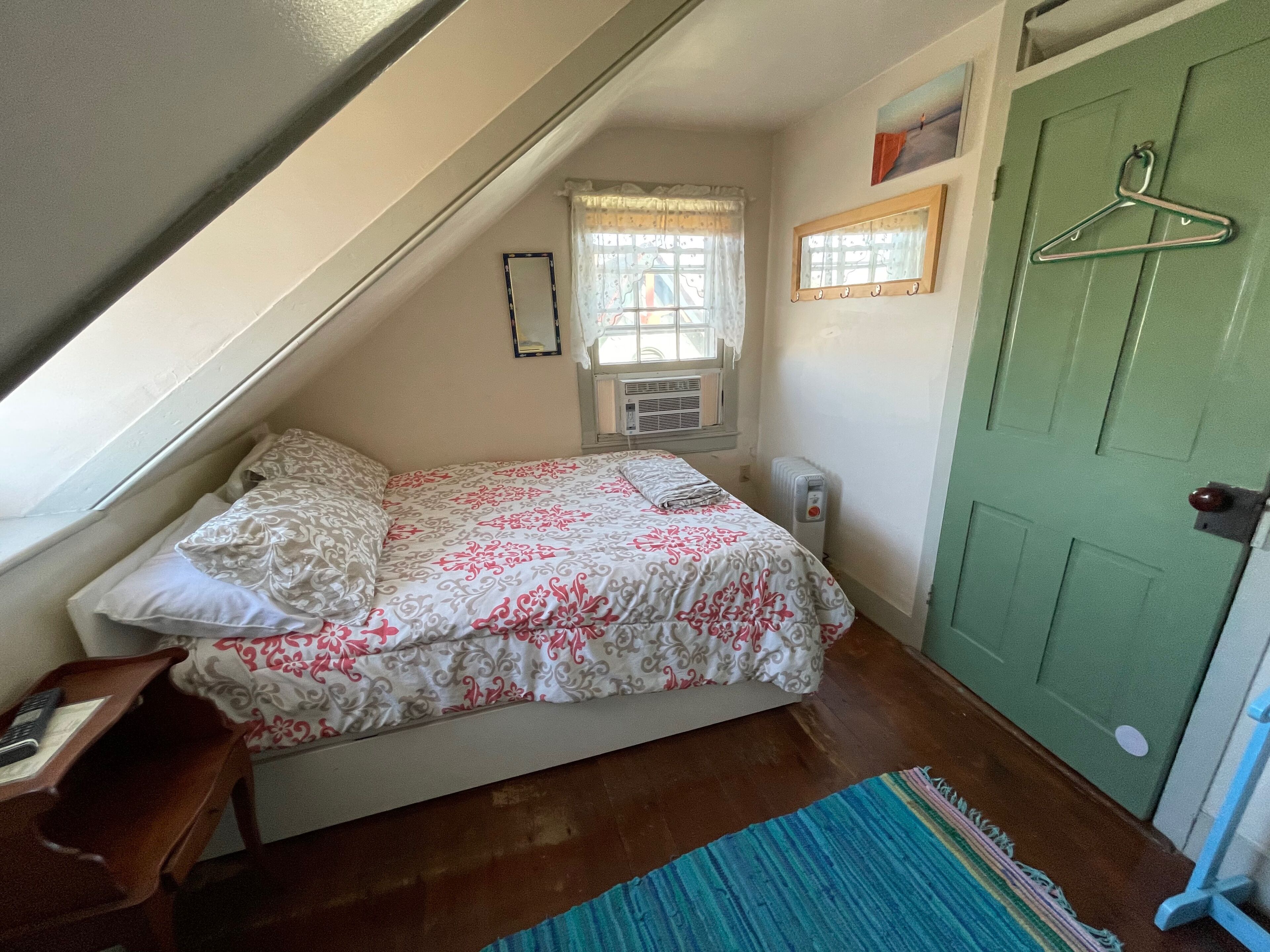 Een kleine, knusse slaapkamer met een schuine kap, een bed met een bloemenbedekking, een raam met gordijn, een radiator en een groene deur.