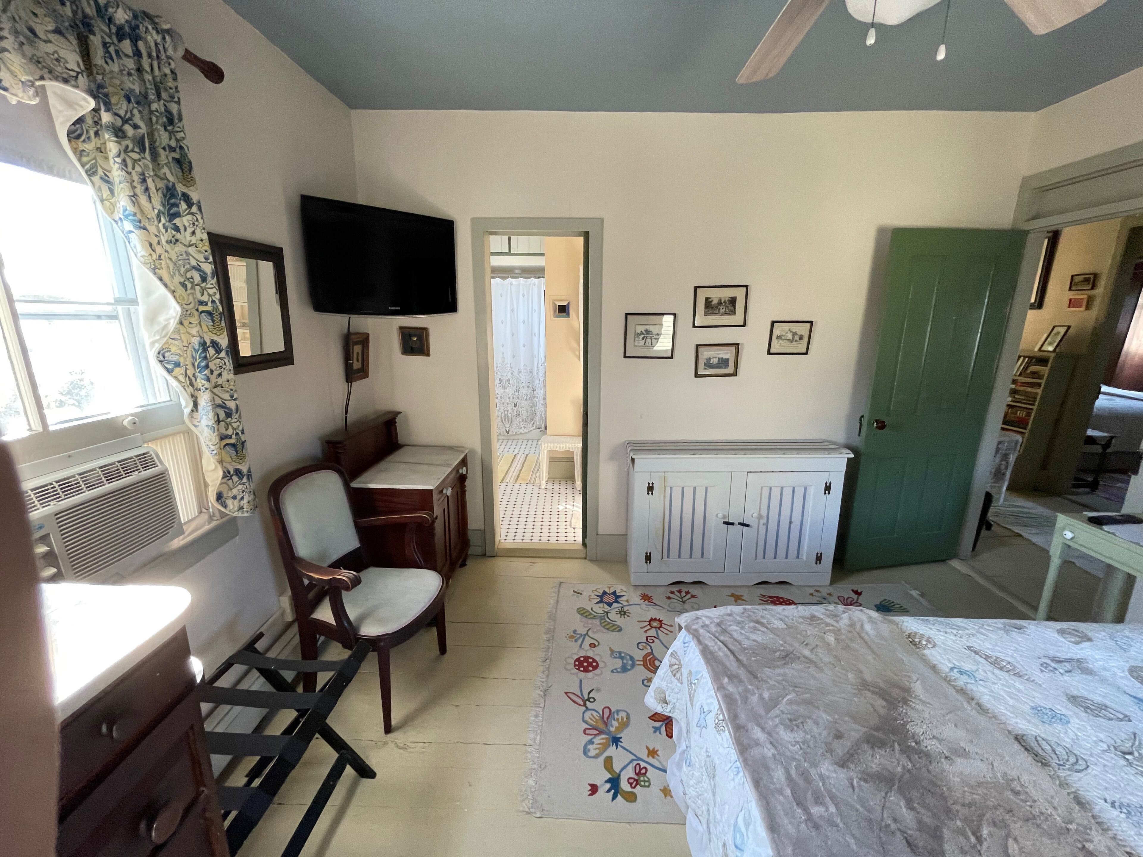 Un dormitorio con cama, escritorio, silla, televisor, radiador y puerta que da a otra habitación.