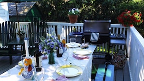 Un área de comedor al aire libre con una mesa dispuesta para cuatro personas, rodeada de sillas, una parrilla y plantas en macetas.