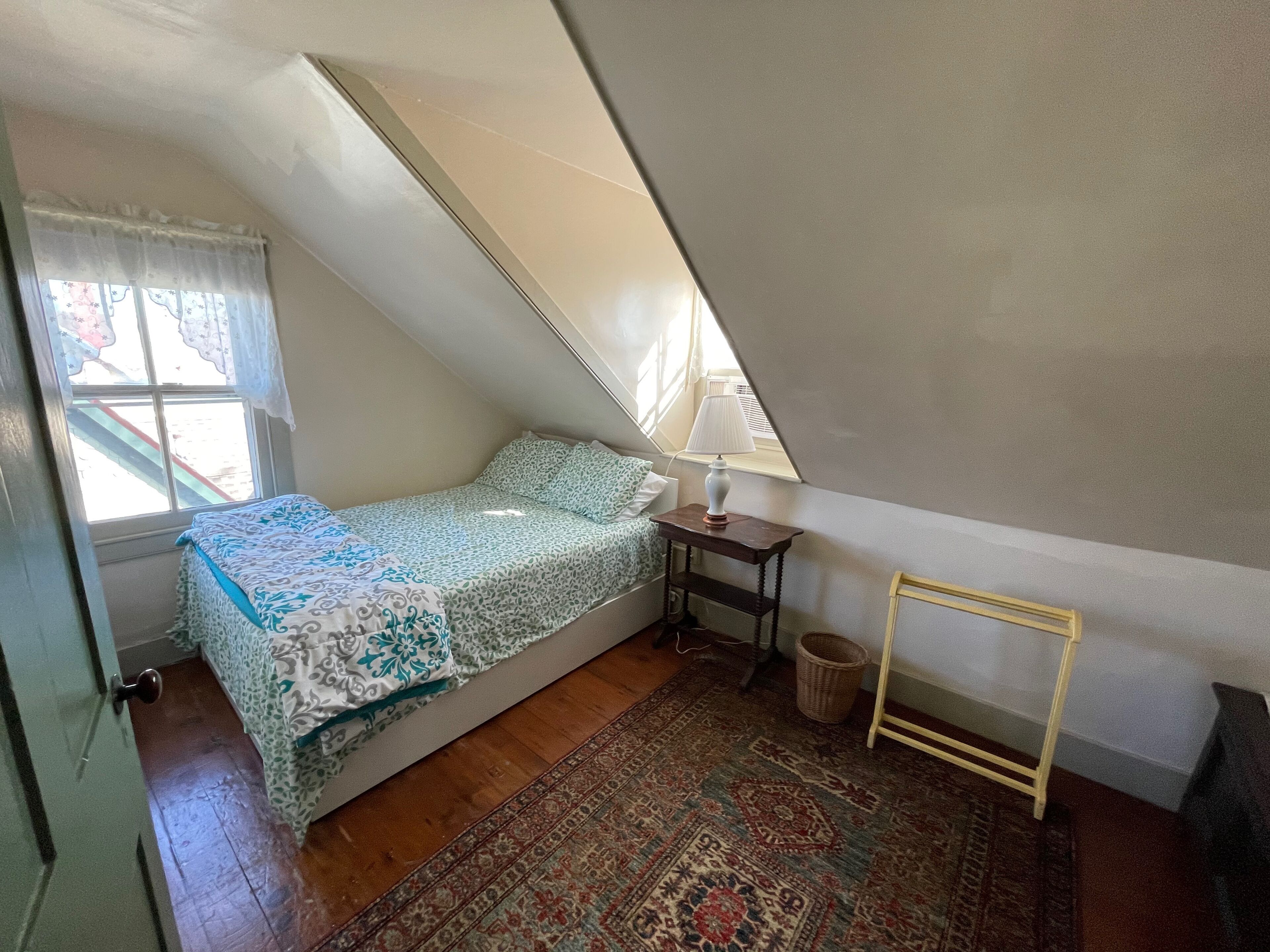 A small, sloped room with a bed, a lamp, a rug, and a window with curtains.