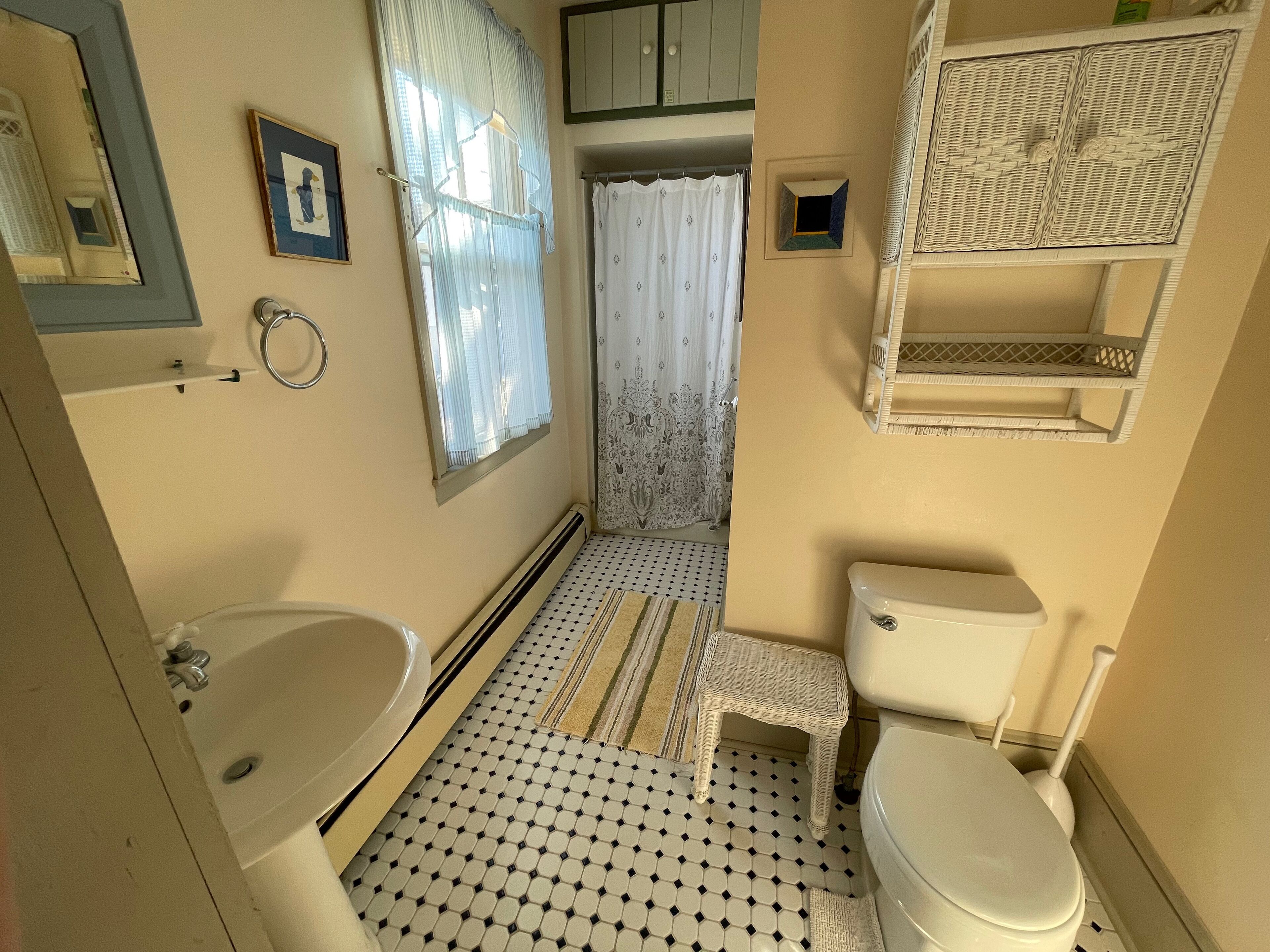 A bathroom with a toilet, sink, and shower area. There is a small white stool and a wicker shelf above the toilet.