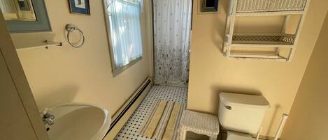 A bathroom with a toilet, sink, and shower area. There is a small white stool and a wicker shelf above the toilet.