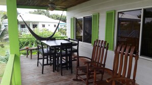 A wooden deck with a table and chairs, a hammock, and a view of a white building with green shutters. - Bungalow 15 - Beachfront Pool View Private Gem! (Playa Hermosa)