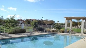 Ein Swimmingpool mit Liegestühlen und einer Pergola, umgeben von einem Zaun und Grünpflanzen.