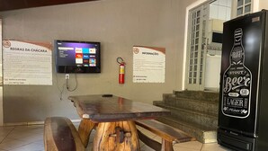 A wooden table with benches, a mounted television, and a refrigerator with a beer advertisement. - Chácara Inribas accommodation and events - Goiânia - GO. (Goiânia)