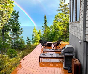 A wooden deck with two chairs and a table, overlooking a forest and a body of water with a rainbow. - Cliff-perched: stunning ocean views, private hiking trails, complete seclusion (Machiasport)