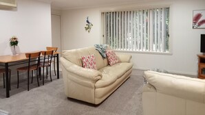 A living room with a beige sofa, two patterned throw pillows, a wooden dining table with chairs, a ceiling fan, and a window with vertical blinds. - The Bottlebrush, 2 bedroom, Villa Unit, Townhouse (Tumut)