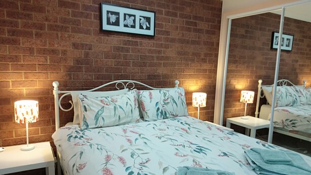 A bedroom with a brick wall, a bed with floral bedding, two bedside lamps, and a mirror. - The Bottlebrush, 2 bedroom, Villa Unit, Townhouse (Tumut)