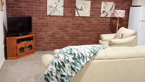 A living room with a brick wall, a flat-screen TV on a wooden stand, a beige sofa, and a ceiling fan. - The Bottlebrush, 2 bedroom, Villa Unit, Townhouse (Tumut)