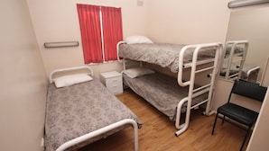 A dormitory room with two bunk beds, a small bedside table, a chair, and a window with red curtains.