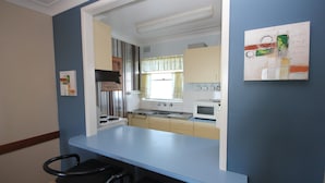 A compact kitchen with a stove, sink, and microwave. There is a window with curtains, and a small dining area with a chair.