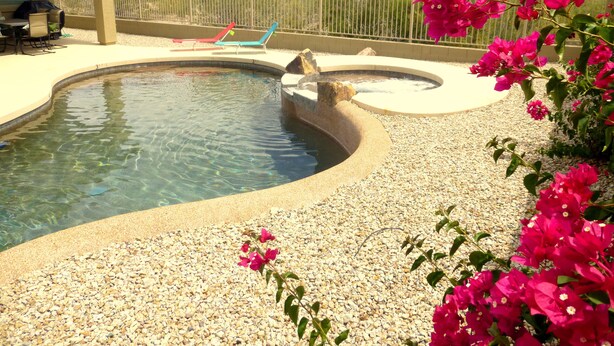 A swimming pool with a pebble surround, lounge chairs, and a covered patio area.