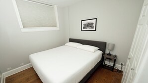 A bedroom with a bed, a nightstand with a lamp, a framed picture on the wall, and a window with blinds. - Wrigley Field All Star Suite 3 Bedrooms 2 Bath (Chicago)