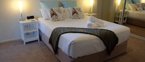 A neatly made bed with a white comforter and decorative pillows, a bedside lamp, and a small white nightstand with a power outlet.