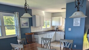 A dining area with a wooden table and chairs, a kitchen in the background, and a sign on the wall.