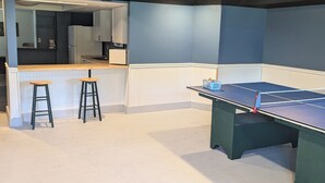 A room with a ping pong table, two stools, a kitchen area, and a ceiling with exposed beams.