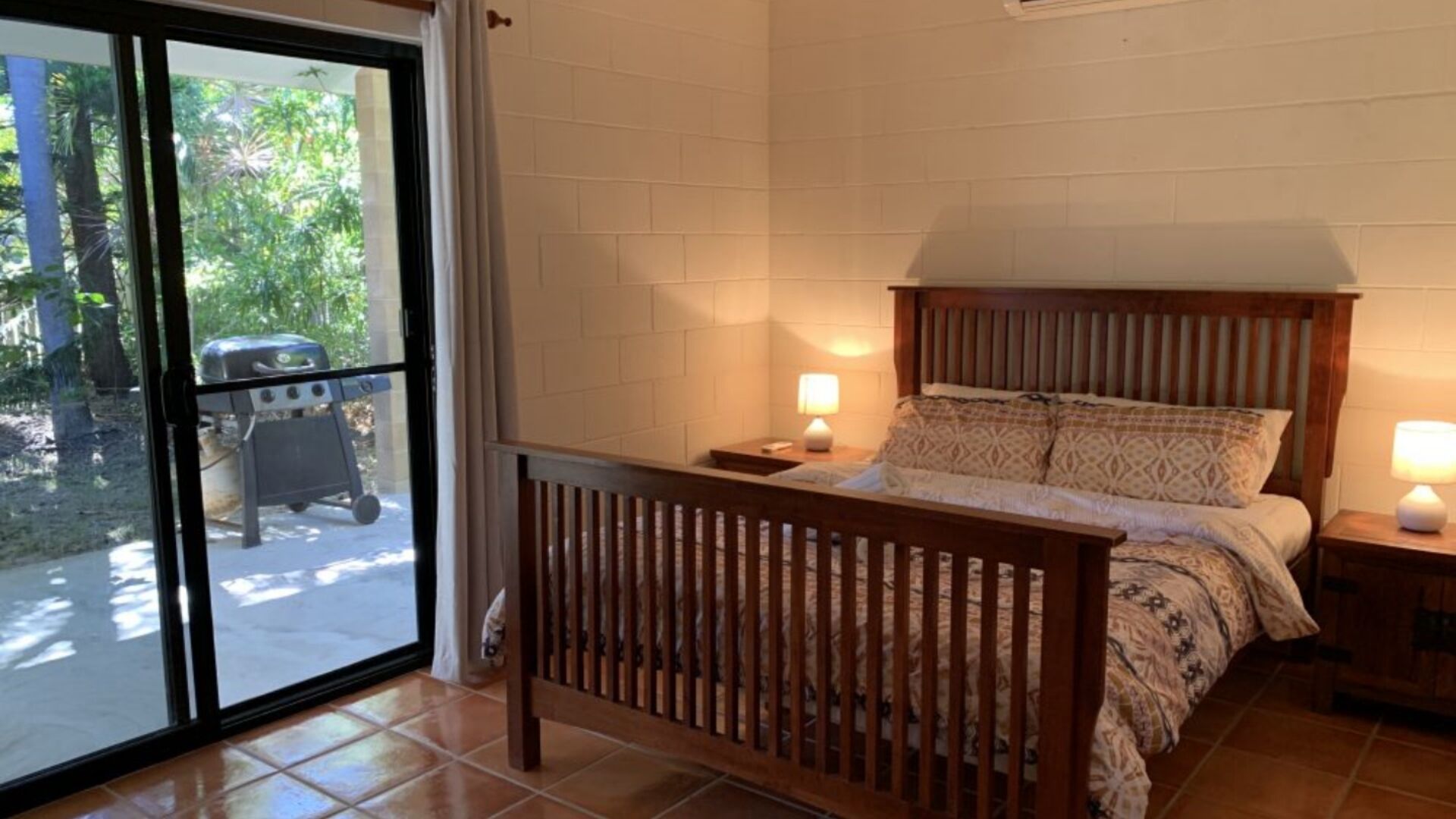 A bedroom with a wooden headboard, a bed with patterned bedding, two bedside lamps, and a view of a barbecue grill outside.