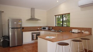 Eine Küche mit einem Edelstahl-Kühlschrank, einer Mikrowelle, einem Backofen, einer Spüle und einem Fenster mit Jalousien. An der Kochinsel stehen drei Hocker.