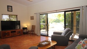 Ein Wohnzimmer mit Fernseher, einer Couch, einem Sessel und einem Tisch. Eine Glastür führt zu einem Außenbereich.