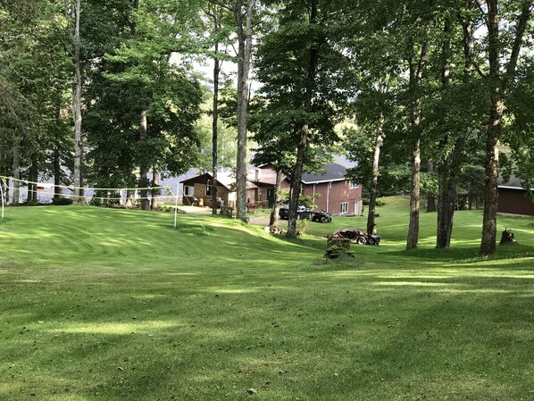 Une zone herbeuse bien entretenue, avec un filet de volley-ball, entourée d’arbres et bordée d’un bâtiment au toit rouge.