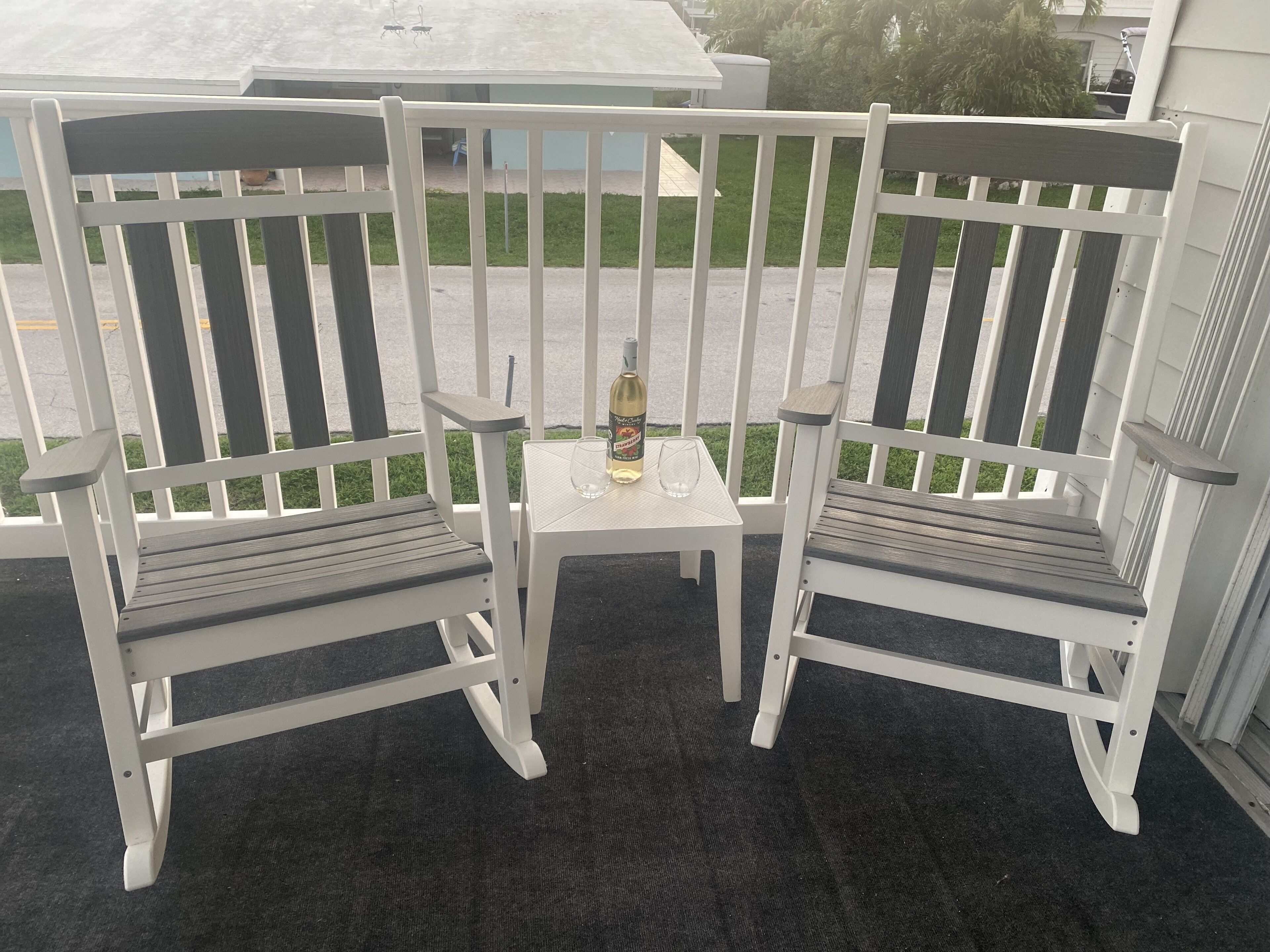 Zwei weiße Holzstühle mit Metallbeinen und ein kleiner Tisch mit Flasche und Glas auf einem Balkon.
