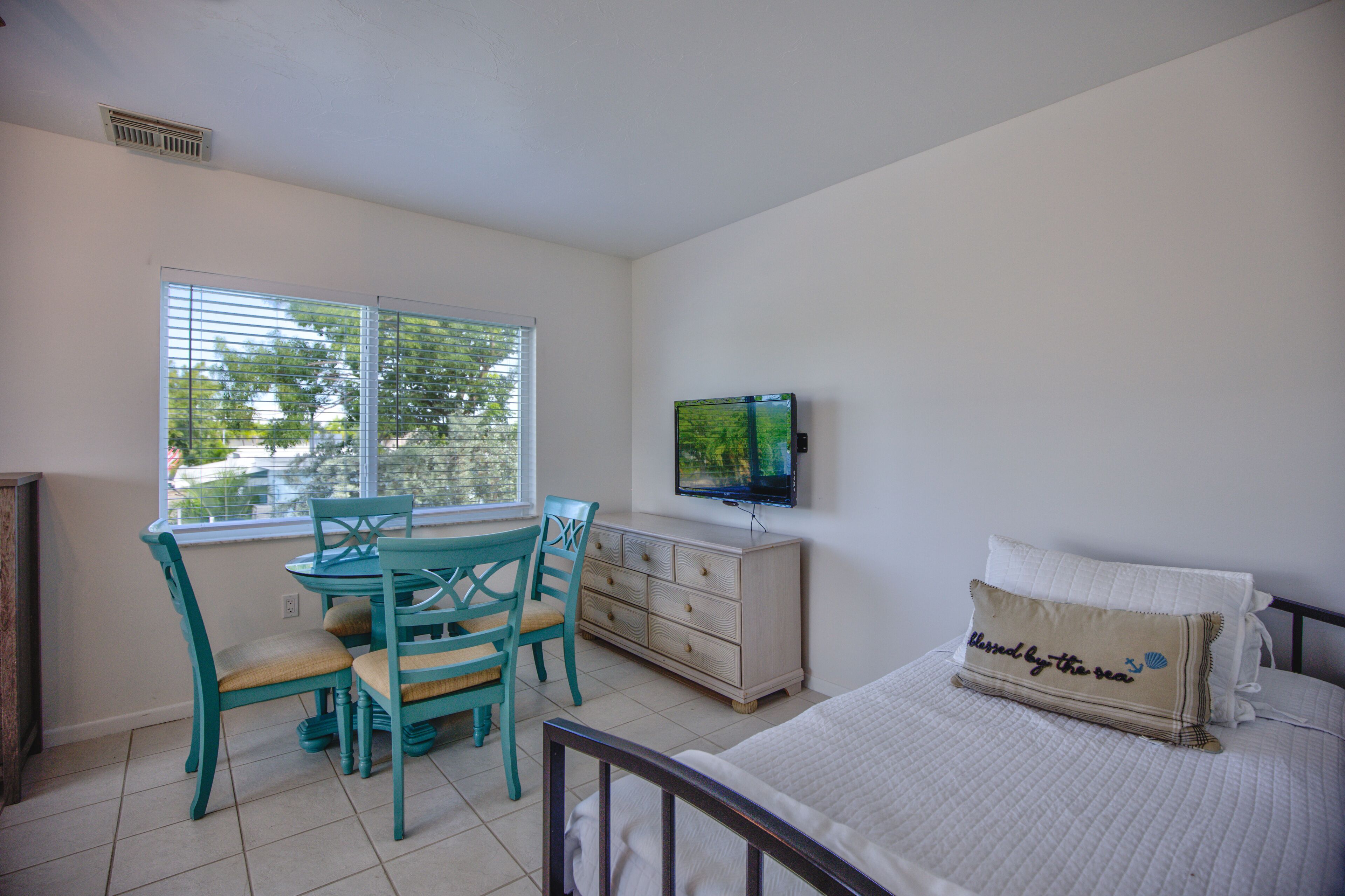 A bedroom with a bed, a TV mounted on the wall, a dresser, and a window with blinds.