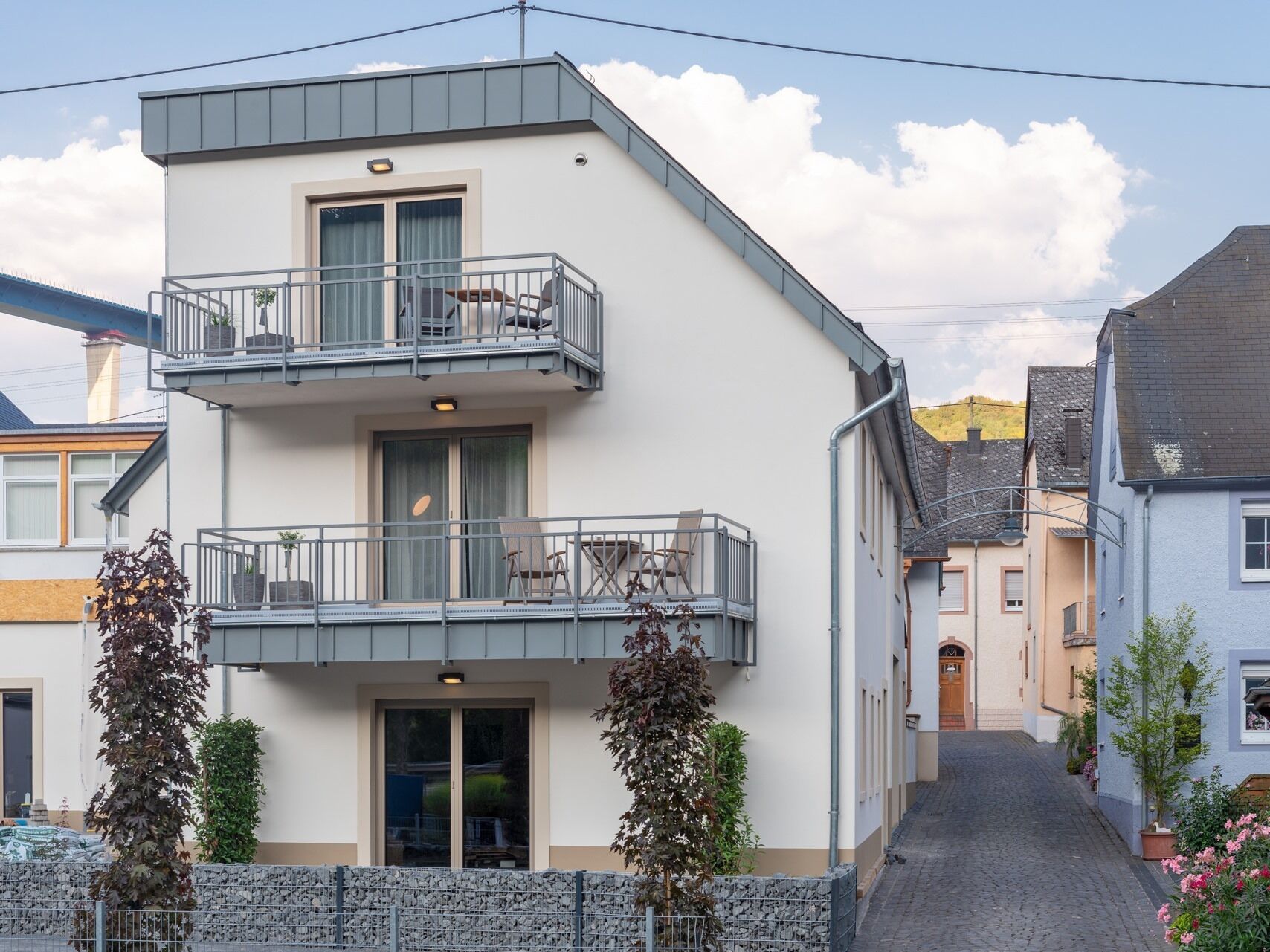 Ein modernes, zweistöckiges Gebäude mit einem Balkon im oberen Stockwerk, einer Steinwand im Vordergrund und einer schmalen Straße im Hintergrund.