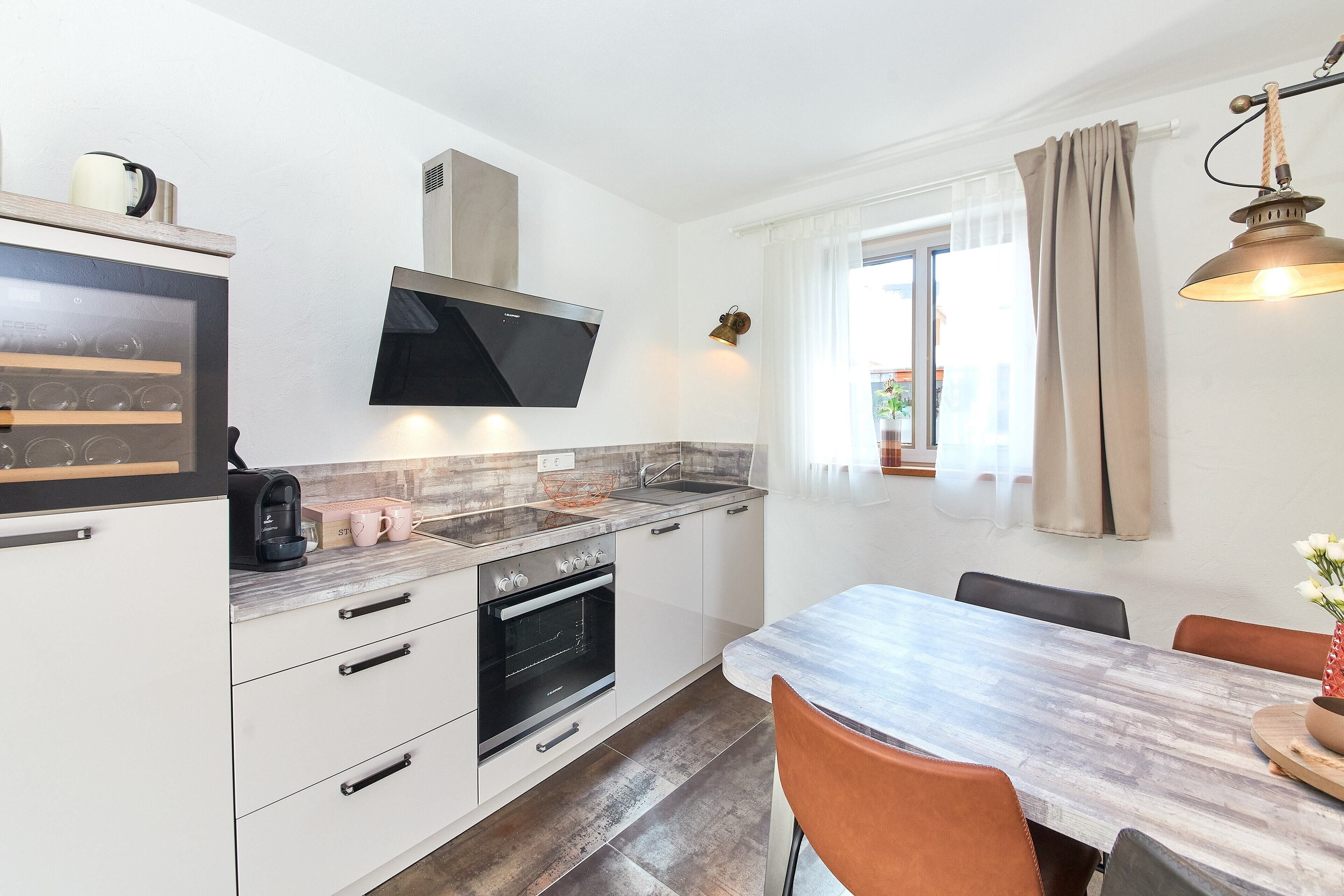 A modern kitchen with white cabinetry, a built-in oven, a microwave, and a coffee maker. There is a dining area with a wooden table and chairs.
