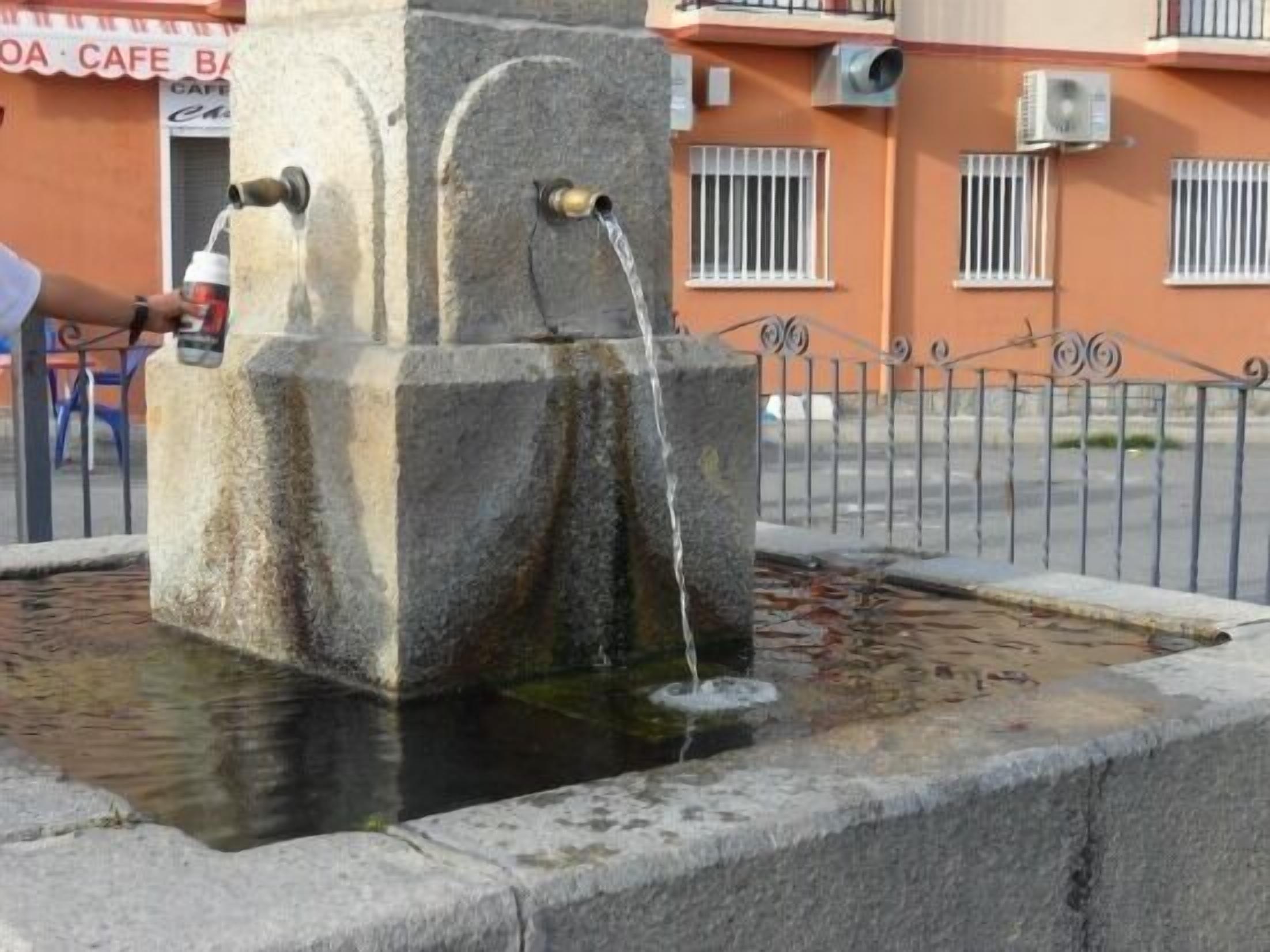 A stone water fountain with two spouts, surrounded by a low stone wall, in front of a building with barred windows and a metal fence.