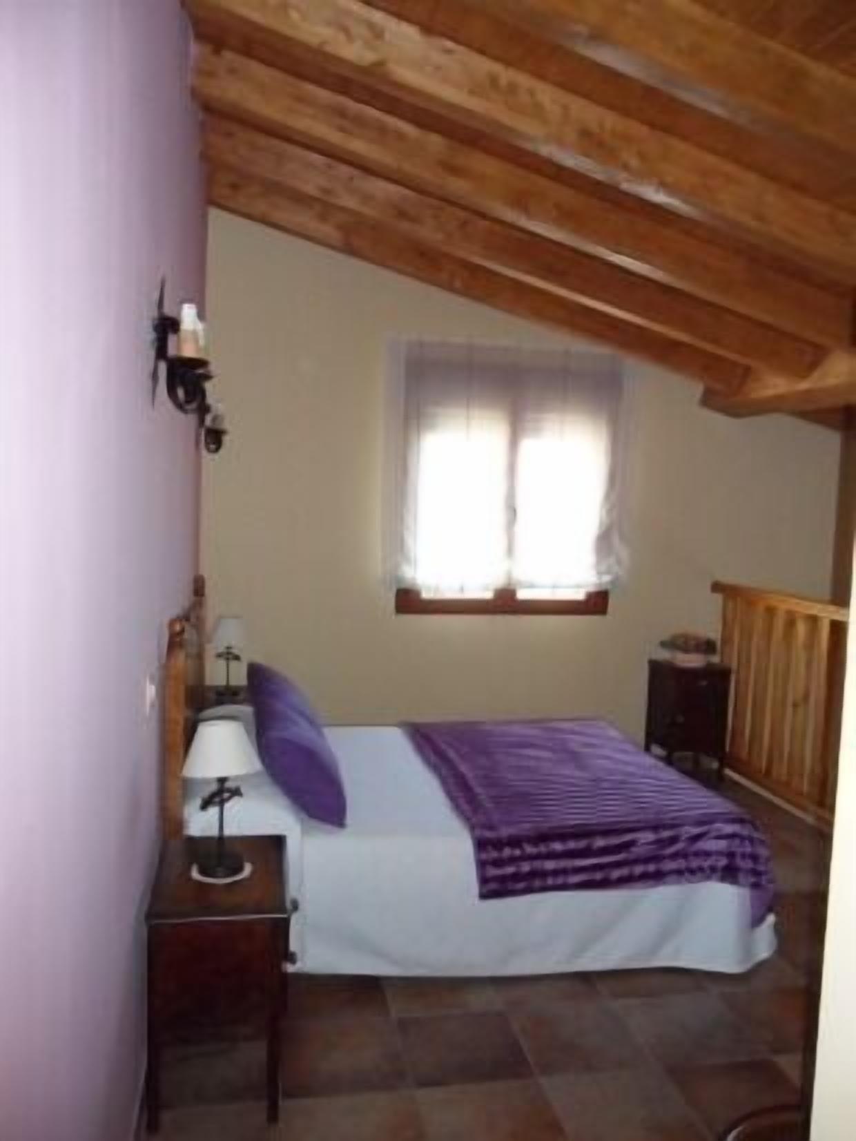 A bedroom with a bed, a nightstand, a lamp, a window, and wooden beams on the ceiling.