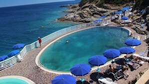 Una piscina a sfioro a forma di ferro di cavallo con acqua cristallina, circondata da lettini e ombrelloni blu, con vista sul mare.