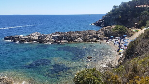 Una spiaggia costiera con formazioni rocciose, acqua cristallina e alcuni ombrelloni a riva.
