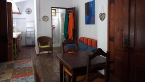 Ein Wohnzimmer mit Holztisch, Sofa und Sessel. Im Hintergrund ist eine Küchenzeile mit Kühlschrank und Spüle zu sehen.