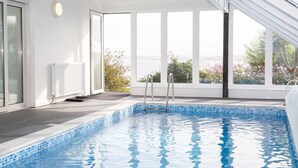 En inomhuspool med kristallklart blått vatten, stora fönster och utsikt över grönområden utanför.