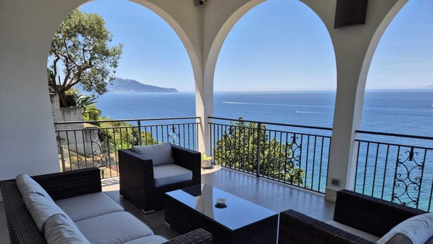 A balcony with wicker furniture, a view of the sea, and a mountain in the distance. - Overview exclusive seaside villa in Sorrento Peninsula (Massa Lubrense Sorrento)
