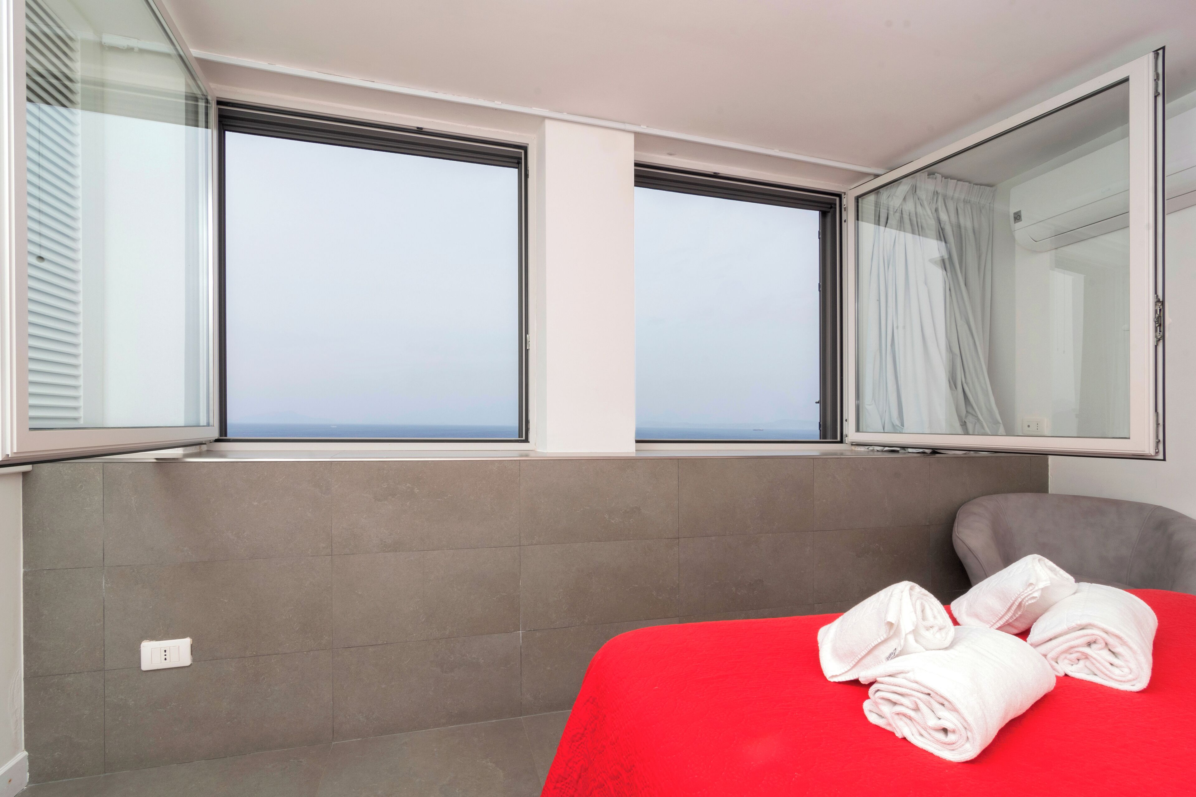 Quarto de hotel com colcha vermelha, duas janelas com cortinas e cabeceira cinza.
